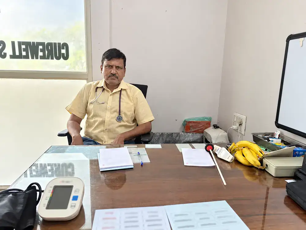 Dr. Mailan K at the desk in the consultation room, with a blood-pressure monitor and an X-ray viewer